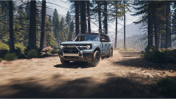 A light blue Ford Bronco Sport driving on a dirt road among pine trees with mountains in the background.